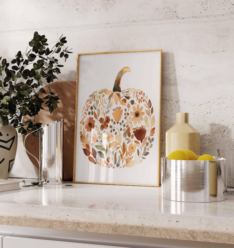 May include: A framed watercolor print of a pumpkin composed of autumn-colored flowers and leaves. The artwork has a gold frame and is displayed on a white countertop. Other decorative items include a vase, a pitcher, and a bowl with lemons.