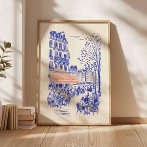 May include: A framed art print depicting a Parisian cafe scene. The illustration features blue line art on an off-white background, with an orange awning and people seated at tables. A tree and buildings are also visible.