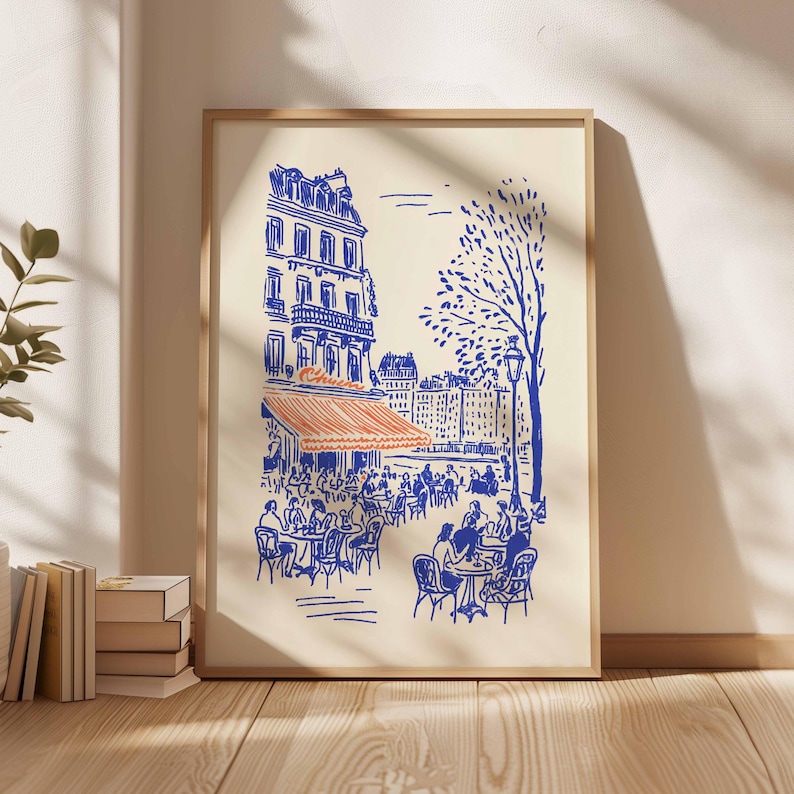 May include: A framed art print depicting a Parisian cafe scene. The illustration features blue line art on an off-white background, with an orange awning and people seated at tables. A tree and buildings are also visible.