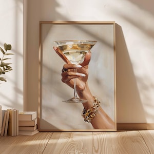 Op de afbeelding: Een ingelijste print van een hand die een martiniglas vasthoudt gevuld met een gouden vloeistof. De hand is versierd met een gouden armband en ringen en heeft lange, witte nagels. Het kunstwerk heeft een neutrale achtergrond.