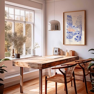 May include: A bright, airy room with a wooden desk and two chairs. A framed print of a Parisian cafe hangs on the wall. A large window offers a view of trees. A silver pendant light hangs above the desk.
