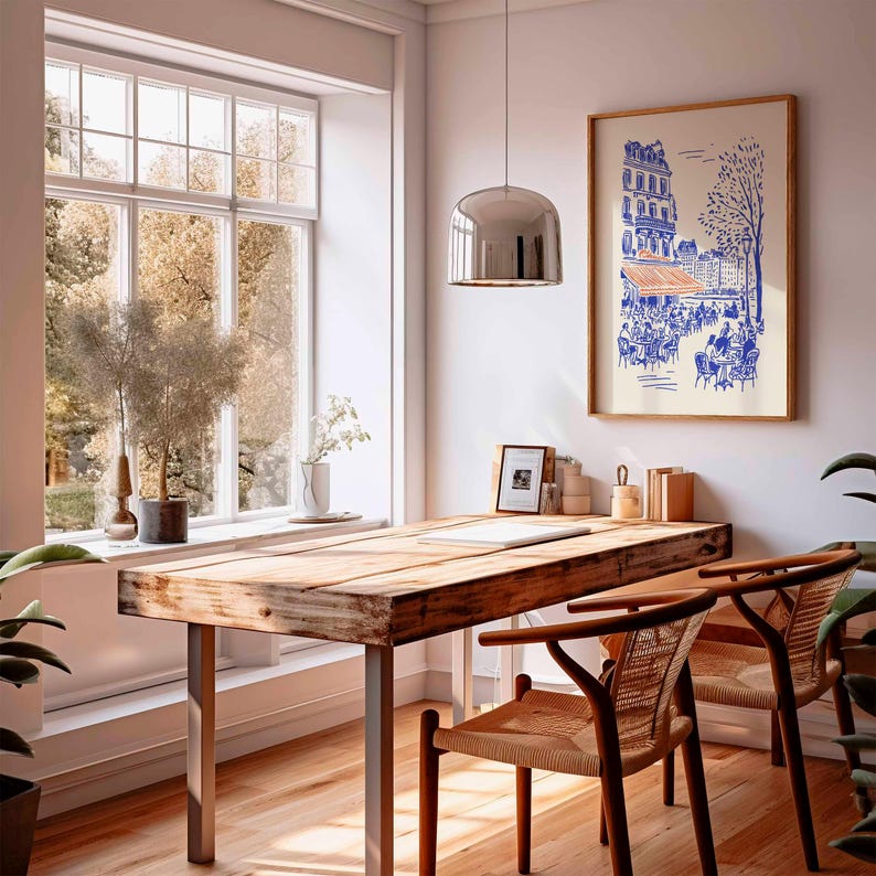 May include: A bright, airy room with a wooden desk and two chairs. A framed print of a Parisian cafe hangs on the wall. A large window offers a view of trees. A silver pendant light hangs above the desk.