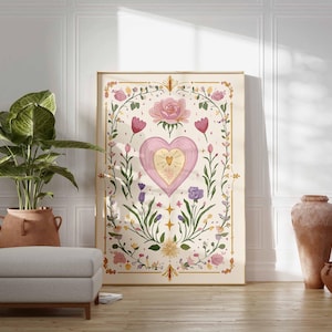 May include: A framed art print featuring a pink heart with a golden center, surrounded by floral and botanical elements. The artwork has a soft, watercolor style with a cream-colored background and gold accents. The print is displayed in a room with a plant, ottoman, and decorative vases.