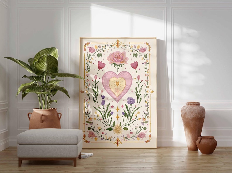 May include: A framed art print featuring a pink heart with a golden center, surrounded by floral and botanical elements. The artwork has a soft, watercolor style with a cream-colored background and gold accents. The print is displayed in a room with a plant, ottoman, and decorative vases.