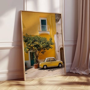 May include: A framed print of a sunny Italian street scene. A yellow building with a small balcony and a lemon tree in front of it. A yellow vintage car is parked on the cobblestone street. The print is leaning against a white wall.