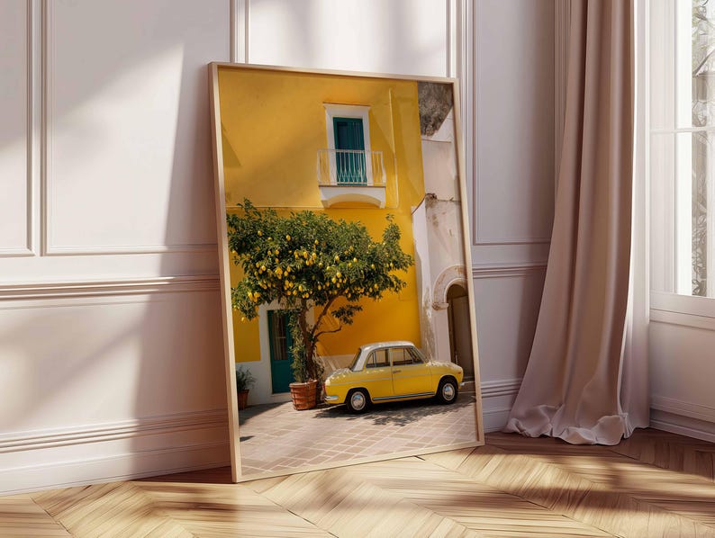May include: A framed print of a sunny Italian street scene. A yellow building with a small balcony and a lemon tree in front of it. A yellow vintage car is parked on the cobblestone street. The print is leaning against a white wall.