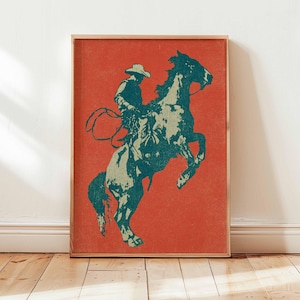 May include: A framed print featuring a teal and off-white illustration of a cowboy on a rearing horse against a red background. The cowboy wears a hat and holds a lasso. The artwork is displayed on a wooden floor.