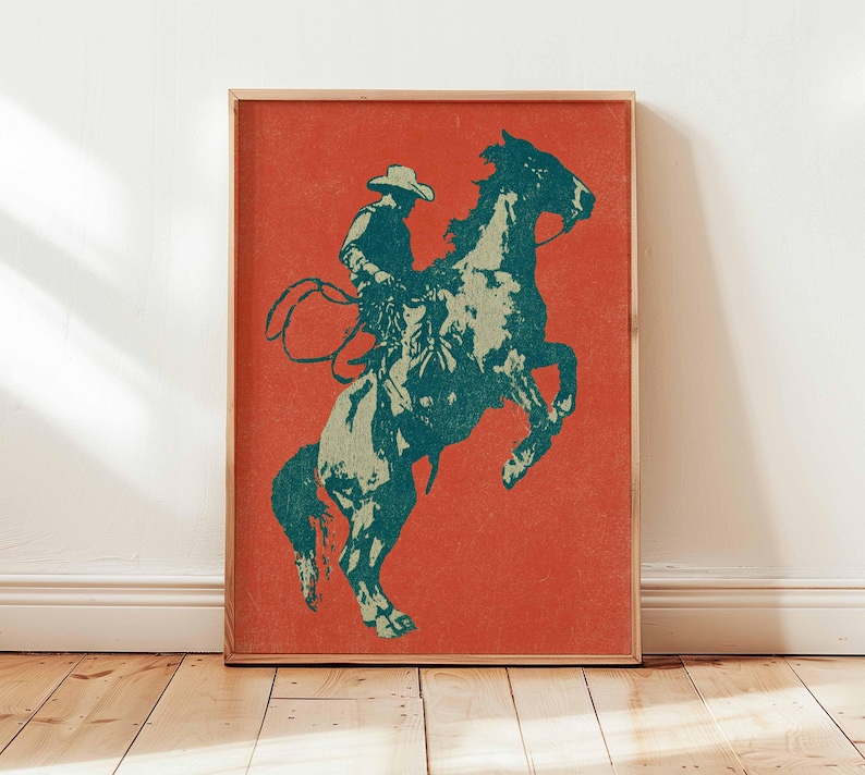 May include: A framed print featuring a teal and off-white illustration of a cowboy on a rearing horse against a red background. The cowboy wears a hat and holds a lasso. The artwork is displayed on a wooden floor.
