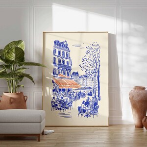 May include: A framed art print depicts a Parisian cafe scene in blue and orange on a cream background. The illustration shows a building, cafe tables, and a tree. The print is displayed with a potted plant, ottoman, and decorative vases.