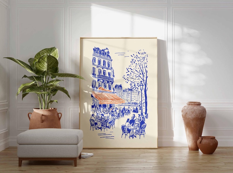 May include: A framed art print depicts a Parisian cafe scene in blue and orange on a cream background. The illustration shows a building, cafe tables, and a tree. The print is displayed with a potted plant, ottoman, and decorative vases.