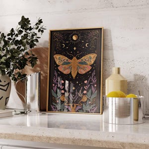 May include: Framed art print featuring a detailed moth illustration with a black background, gold frame, and celestial elements. The moth has intricate wing patterns in shades of orange, yellow, and green. The artwork includes moons, stars, and floral accents.