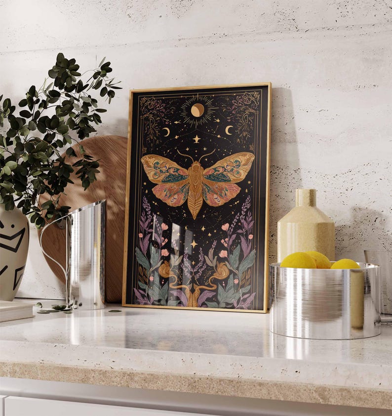 May include: Framed art print featuring a detailed moth illustration with a black background, gold frame, and celestial elements. The moth has intricate wing patterns in shades of orange, yellow, and green. The artwork includes moons, stars, and floral accents.