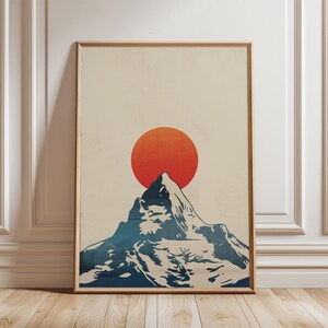 May include: Framed art print depicting a mountain silhouette in blue and white, with an orange sun setting behind it. The artwork has a minimalist style and is set against a cream background. The frame is a light brown colour.
