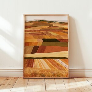 May include: A rectangular framed wall art print featuring a landscape painting of rolling agricultural fields in shades of ochre, golden yellow, deep brown, and olive green, with a distant hill under a pale sky, enclosed in a light wood frame.