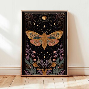 May include: A framed art print featuring a detailed illustration of a moth with intricate wing patterns in shades of gold, pink, and teal against a black background. The artwork includes celestial elements like stars, moons, and suns, with floral and botanical accents.