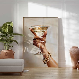 Op de afbeelding: Een kunstafdruk toont een hand die een martini glas vasthoudt gevuld met een gouden vloeistof. De hand is versierd met gouden sieraden en witte nagellak. Het kunstwerk wordt getoond in een kamer met een plant, een poef en decoratieve vazen.