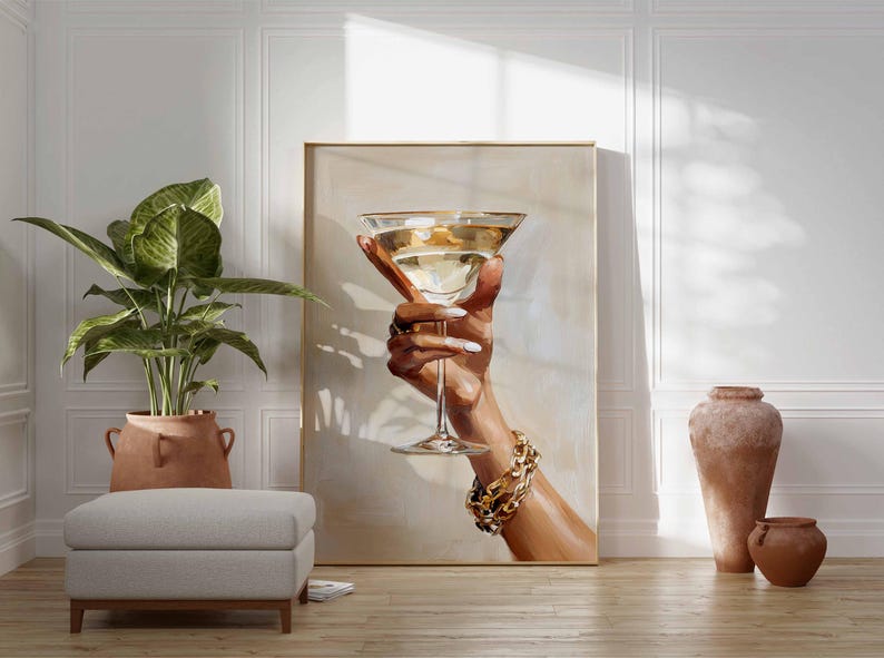 Op de afbeelding: Een kunstafdruk toont een hand die een martini glas vasthoudt gevuld met een gouden vloeistof. De hand is versierd met gouden sieraden en witte nagellak. Het kunstwerk wordt getoond in een kamer met een plant, een poef en decoratieve vazen.