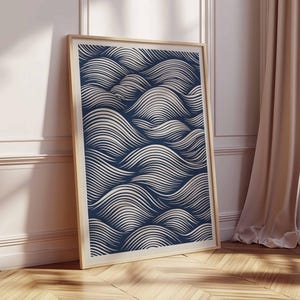 May include: A framed art print featuring a navy blue and white wave pattern. The artwork is displayed in a light wood frame and leans against a white wall and wooden floor. The print's design evokes a sense of movement.