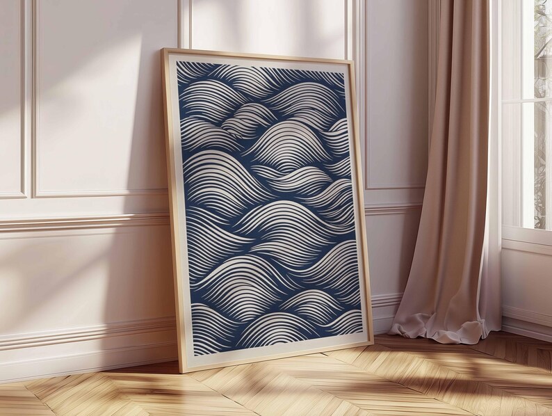 May include: A framed art print featuring a navy blue and white wave pattern. The artwork is displayed in a light wood frame and leans against a white wall and wooden floor. The print's design evokes a sense of movement.