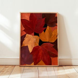 May include: A rectangular framed wall art print featuring a layered composition of autumn leaves in shades of deep red, burnt orange, golden yellow, and dark maroon. The leaves are arranged in an overlapping pattern against a dark background.
