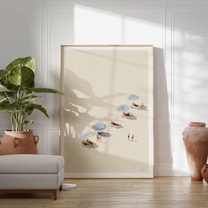 May include: A framed art print showing a beach scene with beach umbrellas and people. The artwork is displayed in a room with a large potted plant, a gray ottoman, and decorative pottery. The color palette is neutral with warm tones.