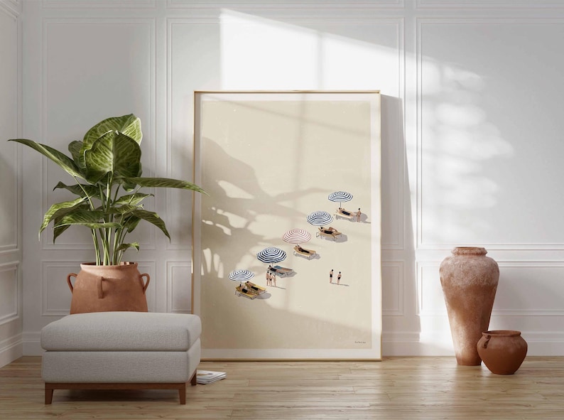May include: A framed art print showing a beach scene with beach umbrellas and people. The artwork is displayed in a room with a large potted plant, a gray ottoman, and decorative pottery. The color palette is neutral with warm tones.
