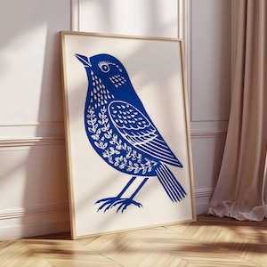 May include: A framed art print featuring a stylized blue bird illustration. The bird is depicted with intricate white floral patterns on its body and wings. The artwork is set against a cream background, with a wooden frame.