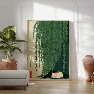 May include: A framed art print featuring a green weeping willow tree with a sleeping white and orange cat beneath its branches. The artwork is displayed in a room with a potted plant, a gray ottoman, and decorative vases.