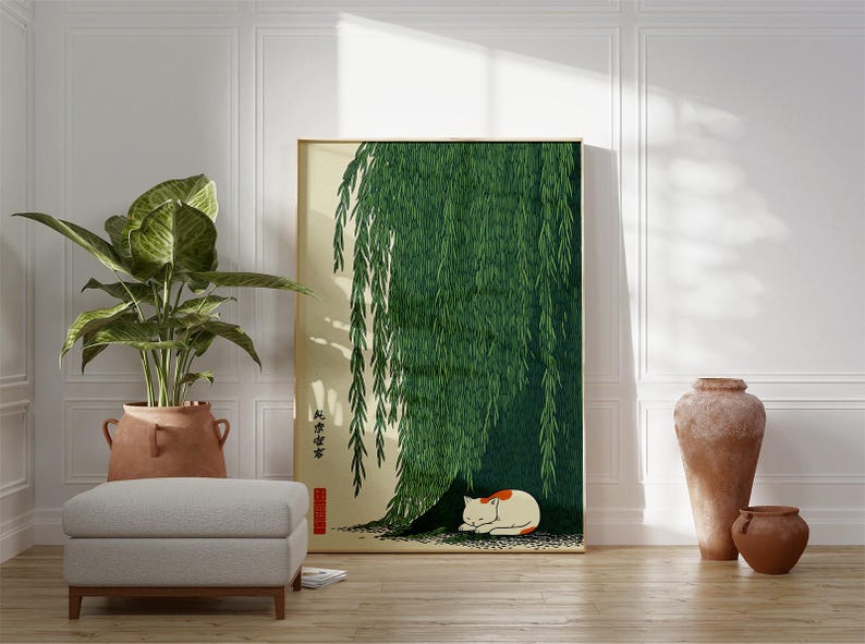 May include: A framed art print featuring a green weeping willow tree with a sleeping white and orange cat beneath its branches. The artwork is displayed in a room with a potted plant, a gray ottoman, and decorative vases.