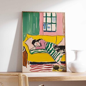 May include: A painting of a woman sleeping on a yellow sofa with a black cat curled up on the blanket. The sofa is in a room with a pink wall and a green curtain. The painting is framed in a wooden frame.