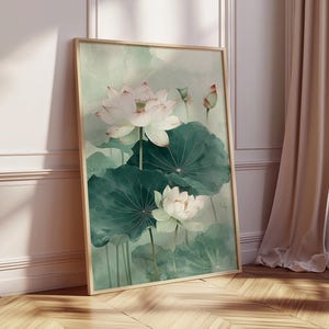 Puede incluir: Una impresi&oacute;n en acuarela enmarcada con flores de loto blancas y hojas verdes. La obra de arte tiene una paleta de colores suaves y apagados con un marco de madera clara. La impresi&oacute;n est&aacute; apoyada contra una pared.