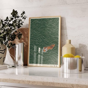 May include: A framed art print featuring a tiger swimming in green water with white wave patterns. The tiger has orange and black stripes. The print is in a wooden frame and is displayed on a white countertop.