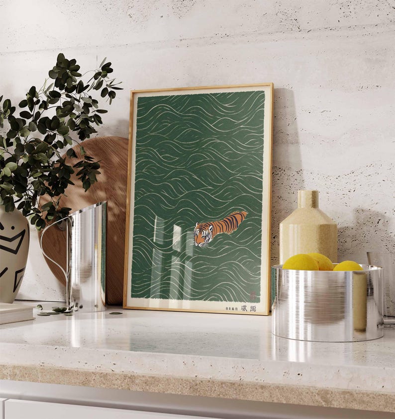 May include: A framed art print featuring a tiger swimming in green water with white wave patterns. The tiger has orange and black stripes. The print is in a wooden frame and is displayed on a white countertop.