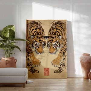 May include: A framed artwork featuring two tigers facing each other, with orange and black stripes and red eyes. The artwork has a gold background with black floral accents and a red seal. The artwork is displayed in a room with a plant, ottoman, and vases.
