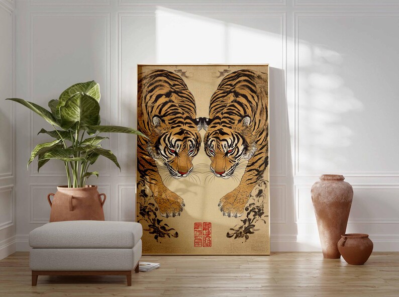 May include: A framed artwork featuring two tigers facing each other, with orange and black stripes and red eyes. The artwork has a gold background with black floral accents and a red seal. The artwork is displayed in a room with a plant, ottoman, and vases.