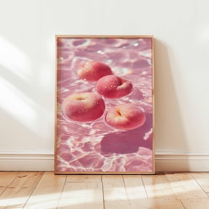 May include: A framed print featuring four pink peaches floating in pink water. The peaches have a slightly flattened shape and are covered in tiny water droplets. The water's surface reflects light, creating a shimmering effect.