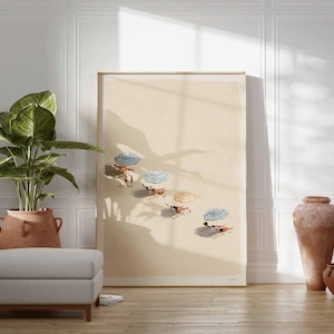May include: A framed art print showing a beach scene with parasols and people sunbathing. The artwork is displayed in a room with a large potted plant, a grey ottoman, and decorative pottery. The room has white panelling and a wooden floor.
