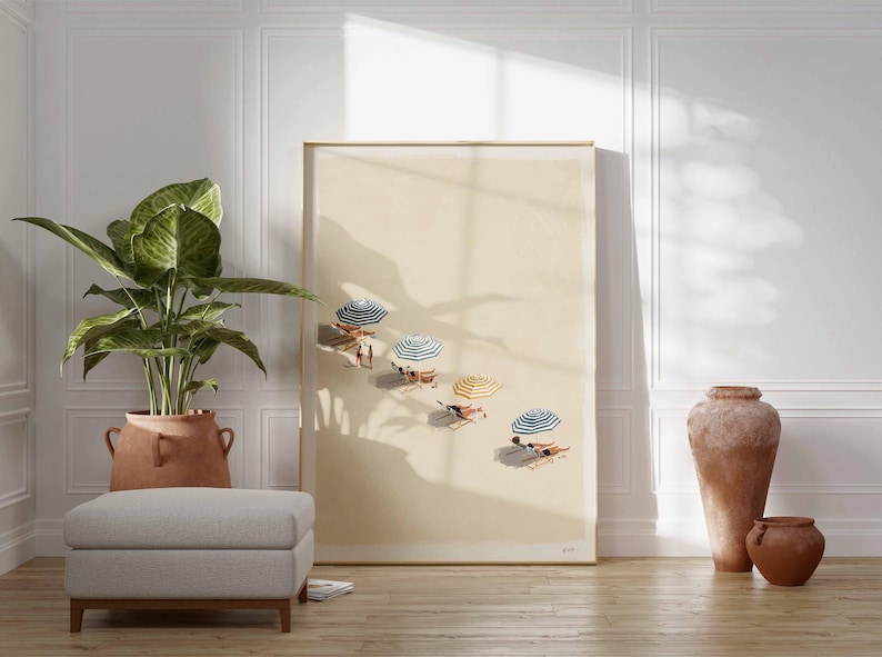 May include: A framed art print showing a beach scene with parasols and people sunbathing. The artwork is displayed in a room with a large potted plant, a grey ottoman, and decorative pottery. The room has white panelling and a wooden floor.