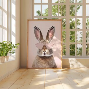 May include: A framed print featuring a brown rabbit wearing pink heart-shaped sunglasses. The rabbit is centered against a light pink background. The artwork is displayed in a room with natural light and a wooden floor.
