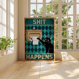 May include: A framed poster with the words "SHIT HAPPENS" and a cartoon image of a black cat reaching for toilet paper. The background is teal with a diamond pattern. The poster is in a room with a window and plants.