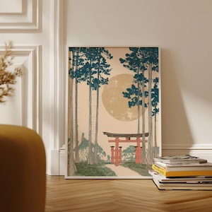 May include: Framed art print featuring a Japanese landscape. The artwork depicts a red torii gate at the end of a path, tall trees with teal foliage, and a large yellow sun. The print is displayed on a wooden floor.