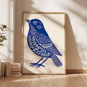 May include: A framed art print featuring a blue bird illustration. The bird is depicted in a linocut style, with intricate details in white. The artwork is set against a cream background, framed in natural wood, and leans against a wall.
