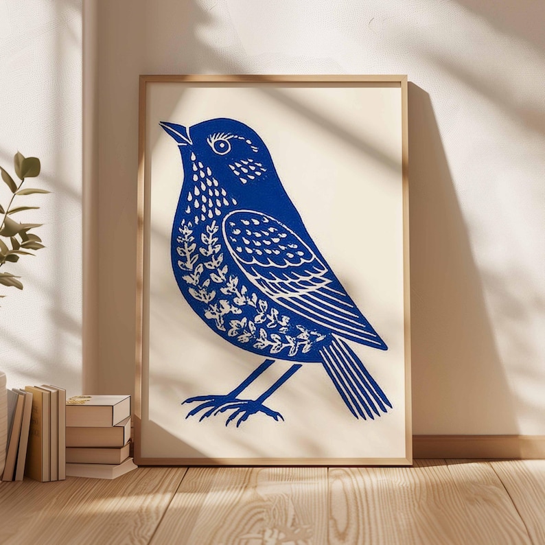 May include: A framed art print featuring a blue bird illustration. The bird is depicted in a linocut style, with intricate details in white. The artwork is set against a cream background, framed in natural wood, and leans against a wall.