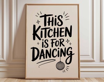 May include: A framed print with the text "THIS KITCHEN IS FOR DANCING" in black script. The print has a beige background and a disco ball illustration. The frame is a light brown color. The print is leaning against a white wall.