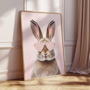 May include: A framed print featuring a close-up of a rabbit wearing pink heart-shaped sunglasses. The rabbit has brown and tan fur with long ears. The background is a soft pink color.