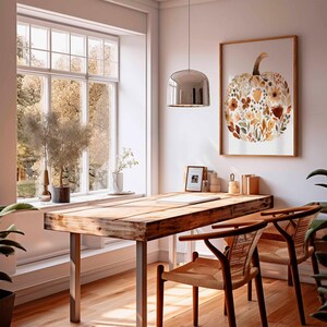 May include: A cozy home office featuring a wooden desk with metal legs, two woven chairs, and a large window. A framed pumpkin art print with floral details hangs on the wall. The room is filled with natural light and plants.