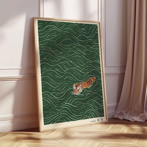 May include: Framed art print featuring a tiger swimming in green water with white wave patterns. The tiger has orange and black stripes. The artwork is in a wooden frame and leans against a white wall and wooden floor.