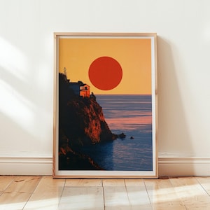 Op de afbeelding: Een ingelijste kunstdruk met een kustlandschap met een grote rode zon tegen een goudgele lucht. Een donkere klif met een klein huis bevindt zich aan de linkerkant en kijkt uit over een kalme blauwe oceaan die het warme licht weerspiegelt.
