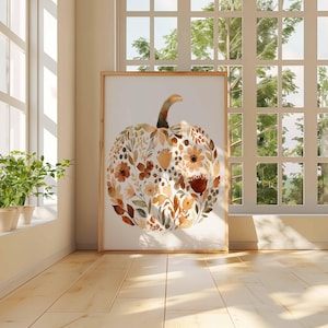 May include: A framed watercolor print of a pumpkin filled with floral elements in shades of brown, orange, and cream. The artwork is displayed in a room with natural light from a large window, creating a warm, inviting atmosphere. The pumpkin art is a fall-themed home decor item.
