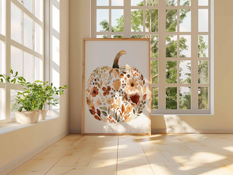 May include: A framed watercolor print of a pumpkin filled with floral elements in shades of brown, orange, and cream. The artwork is displayed in a room with natural light from a large window, creating a warm, inviting atmosphere. The pumpkin art is a fall-themed home decor item.